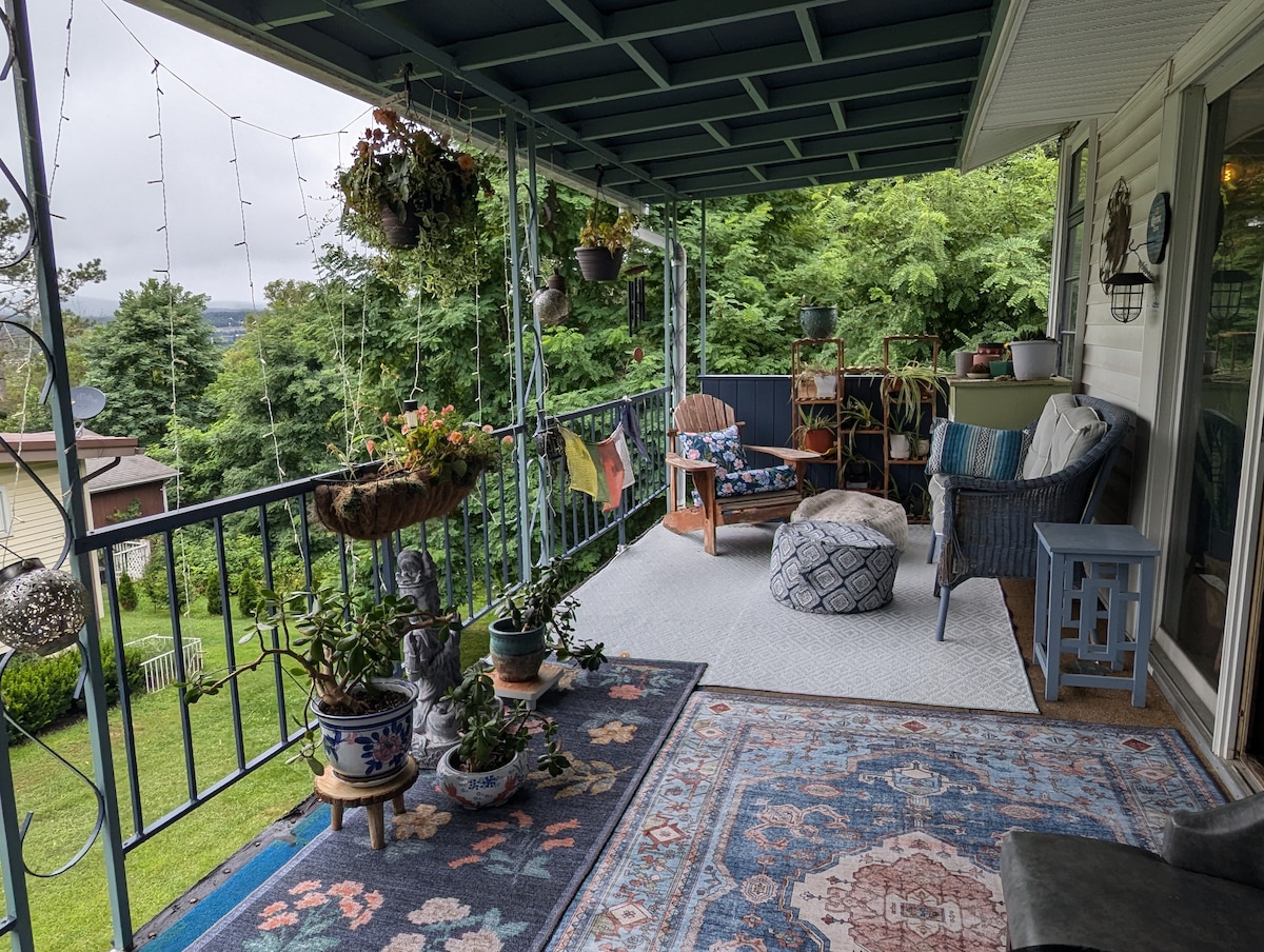 A covered porch is presented with a variety of potted plants and a colorful outdoor rug. Comfortable seating includes a patterned chair and a low table, while hanging planters and string lights add a touch of charm against the green backdrop of trees.