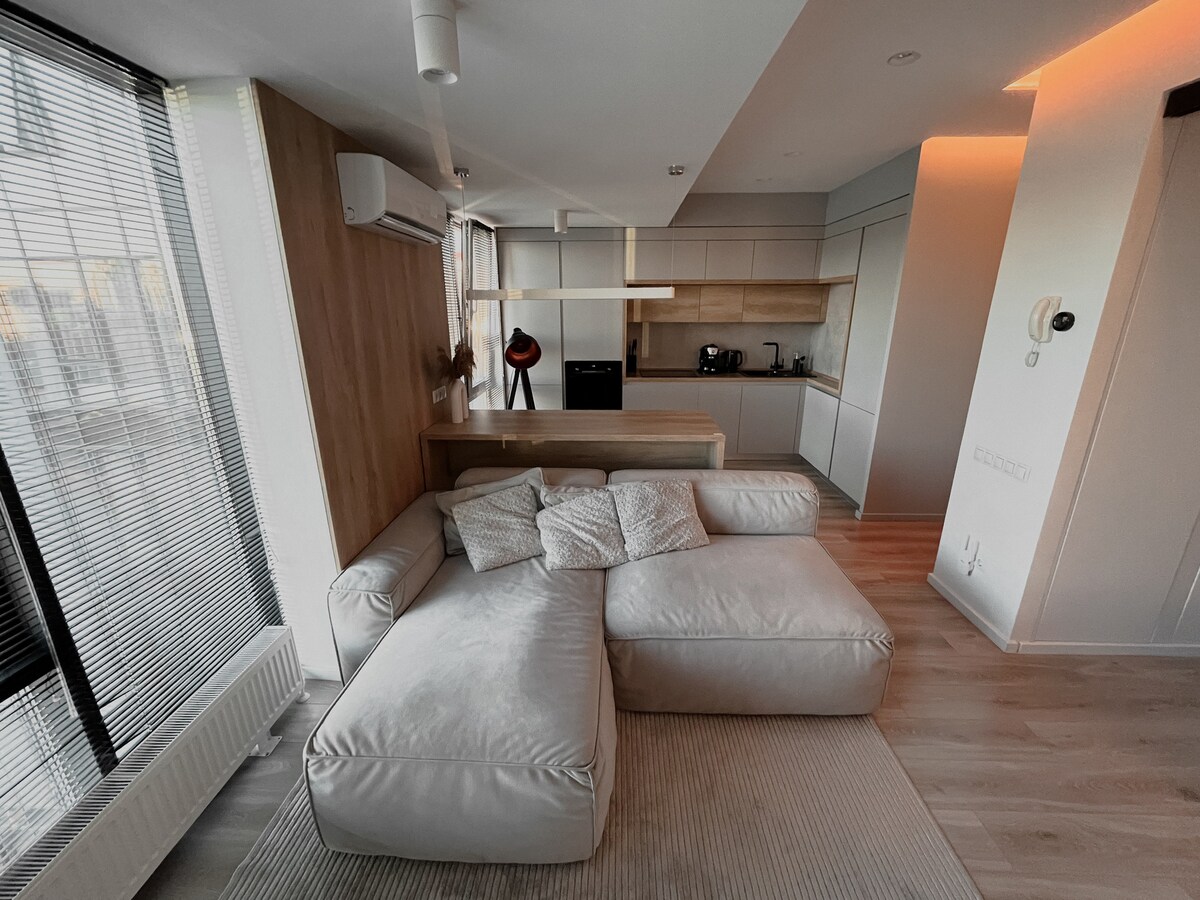 A spacious living area is highlighted by a light-colored sectional sofa arranged on a soft area rug. The modern kitchen features sleek cabinetry and appliances, while large windows allow natural light to fill the room, creating a bright and airy atmosphere.