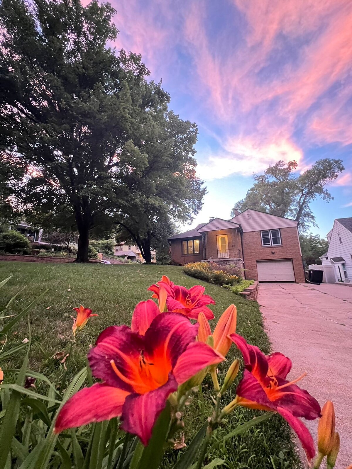 A vibrant garden area features clusters of bright orange and pink flowers, framed by lush green grass. The house stands in the background, showcasing a brick façade, with a garage and large windows under a sky painted with soft hues of blue and pink.