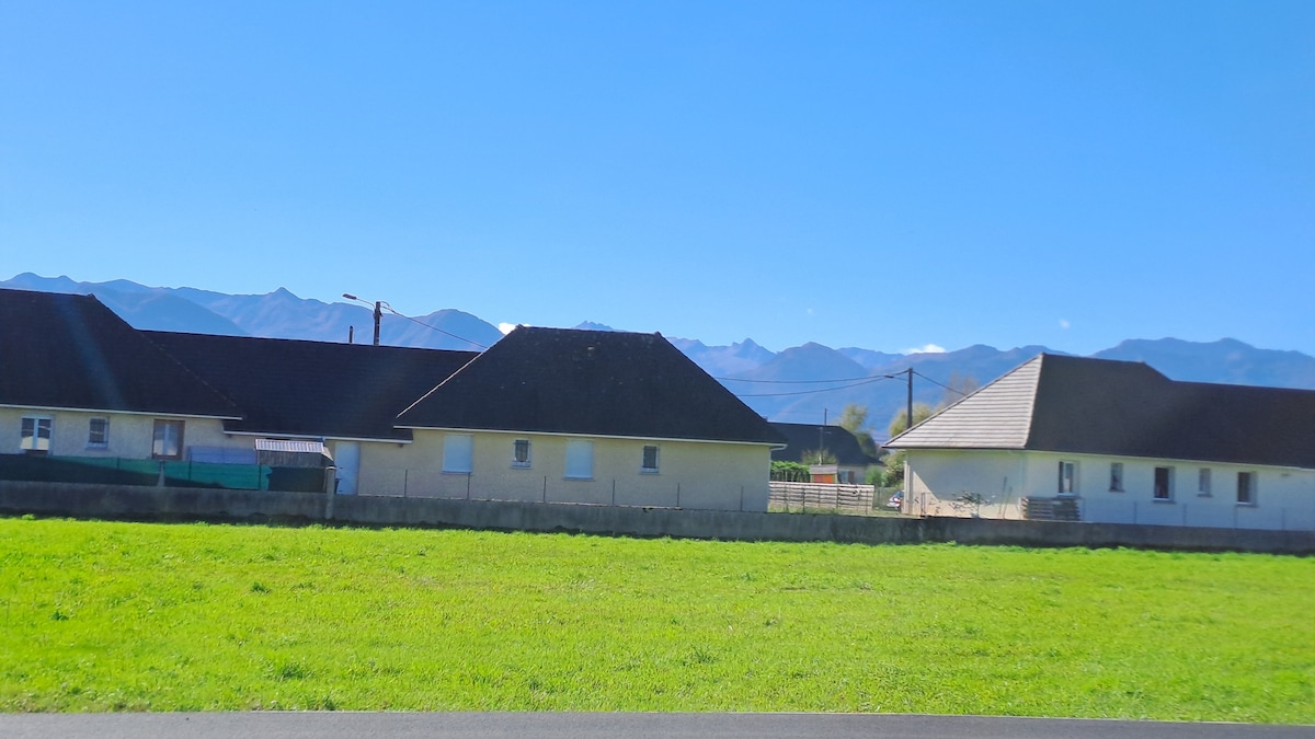 A serene view captures a row of houses set against a backdrop of distant mountains. The landscape features a grassy area in the foreground, with clear blue skies above, emphasizing the peaceful rural setting.