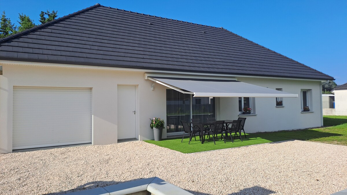 The exterior of a modern one-story house is displayed, featuring a covered patio with an outdoor dining set. The lawn is trimmed, and a garage is visible to the left. A retractable awning provides shade, enhancing comfort for outdoor gatherings.