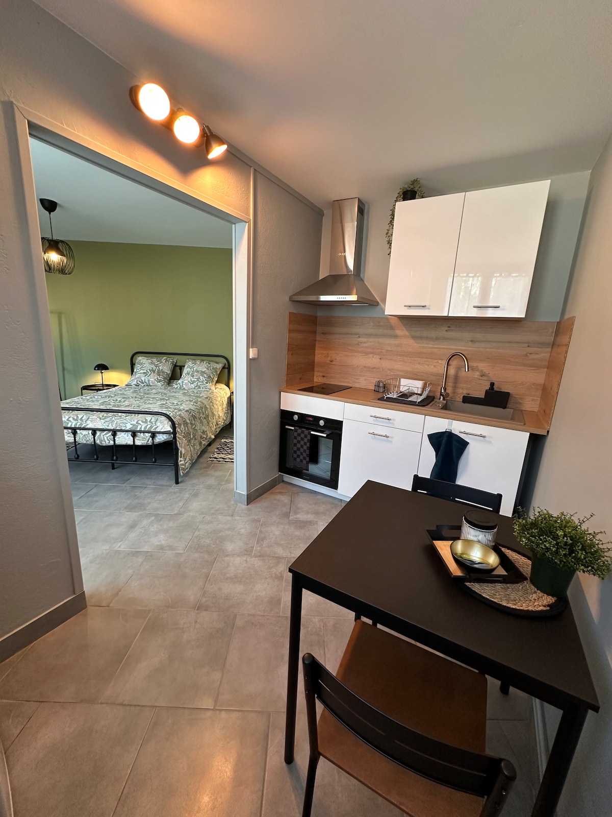 A compact kitchen area features modern cabinetry and essential appliances, including a refrigerator and stovetop. A small dining table with seating is situated nearby, while a glimpse into the living space reveals a spacious bed with tasteful bedding against a green wall.