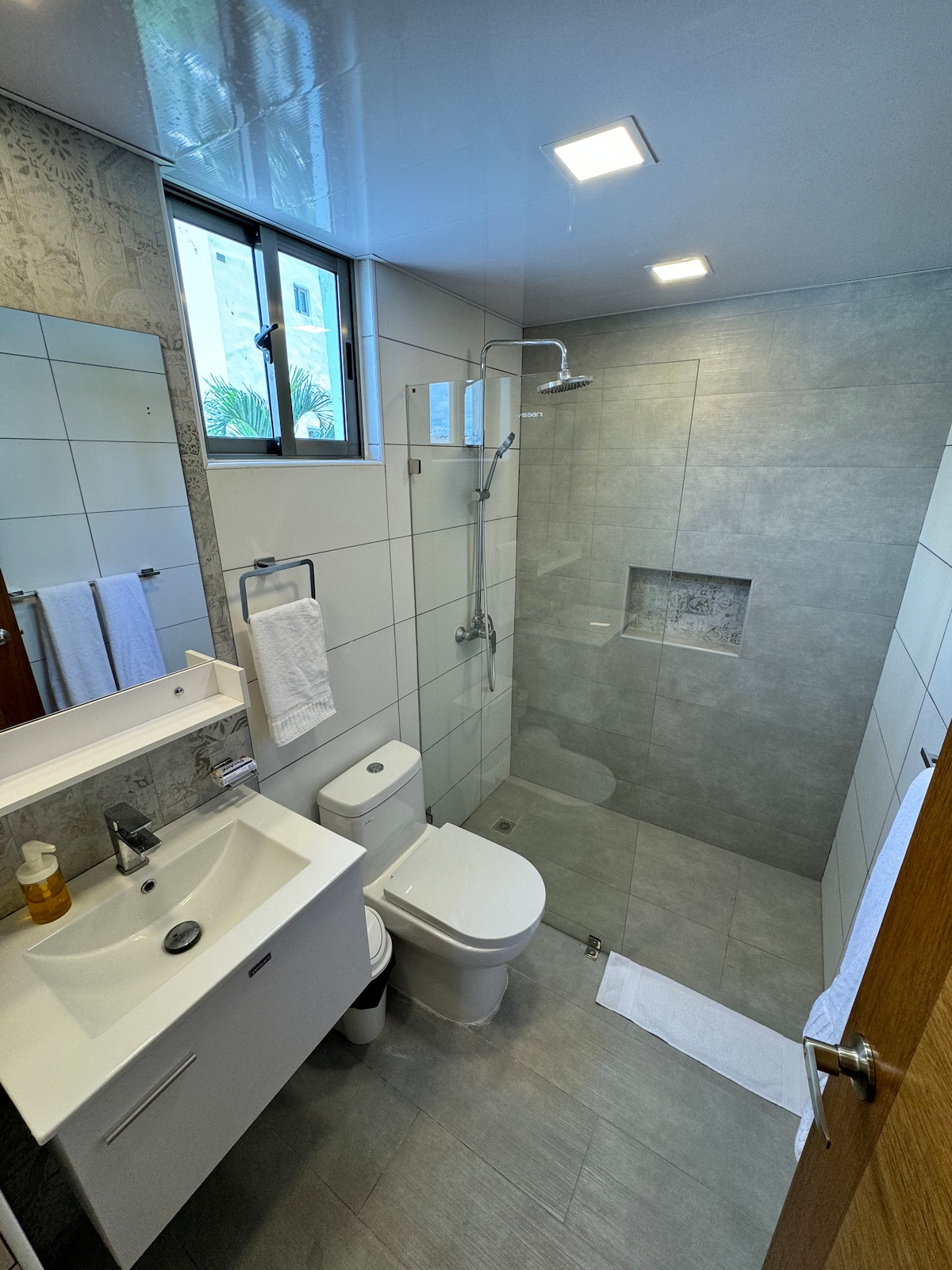 A modern bathroom features a walk-in shower with a glass partition, a sleek vanity with a sink, and a wall-mounted mirror. Towels are neatly arranged, and natural light filters through a window, enhancing the clean, minimalist design of the space.