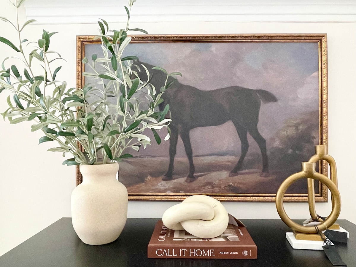 A decorative table display features a vase with greenery beside a sculptural piece. A framed artwork depicting a horse is positioned in the background, while a small stack of books is placed on the table, contributing to the curated aesthetic of the space.