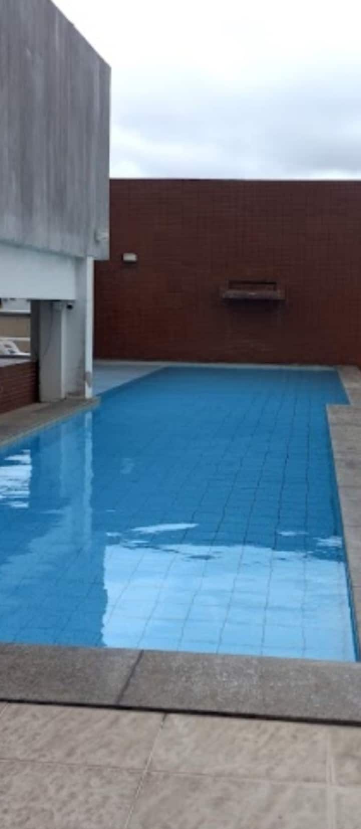 A long, rectangular swimming pool extends alongside a smooth stone surface. The pool features clear, blue water and is bordered by a low wall. A shaded area is visible nearby, enhancing the serene environment.