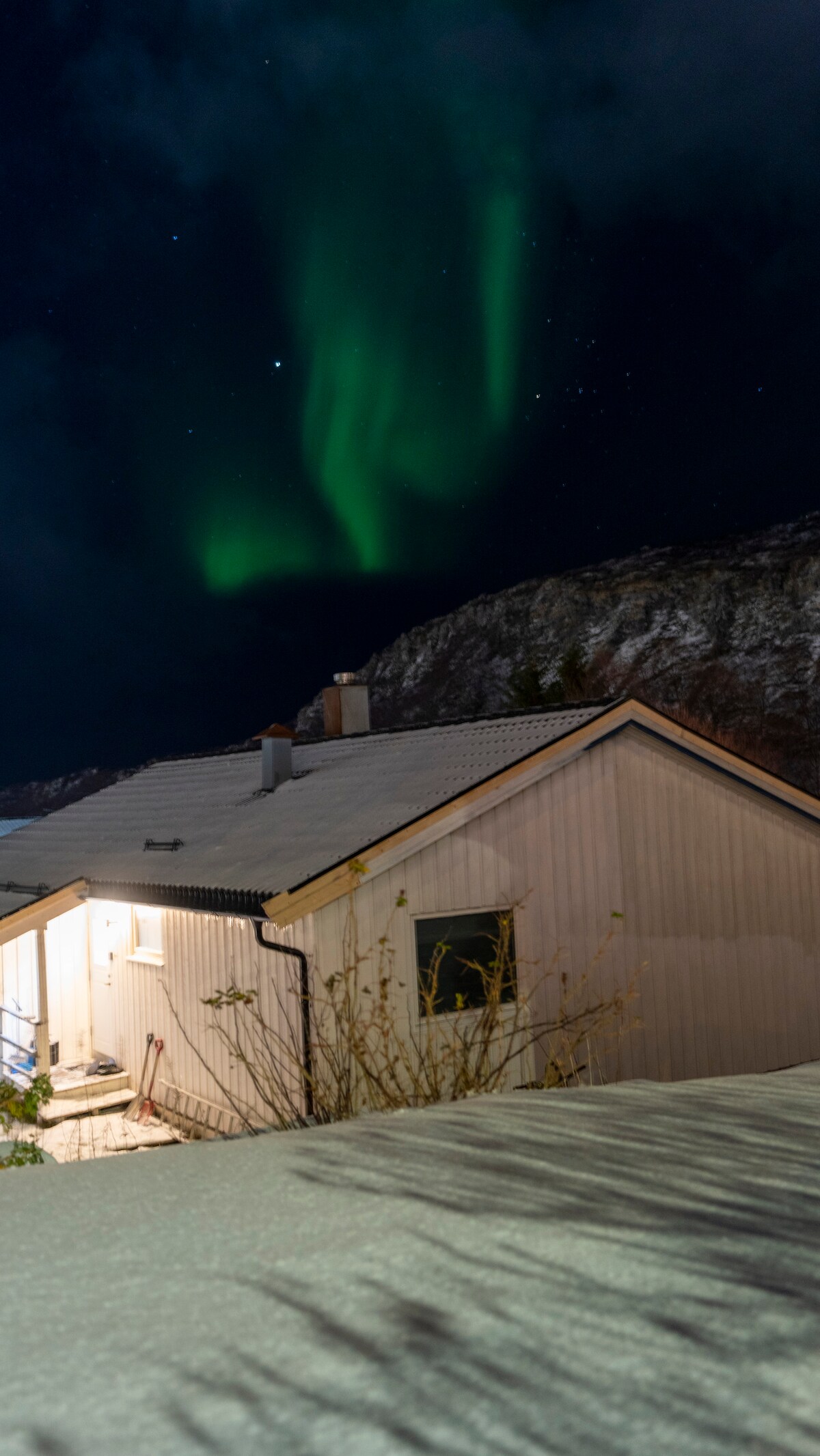 A residential structure is softly illuminated against a backdrop of snow-covered ground and night sky. The northern lights cast a gentle green glow above the home, while a rugged mountainous landscape is visible in the distance.