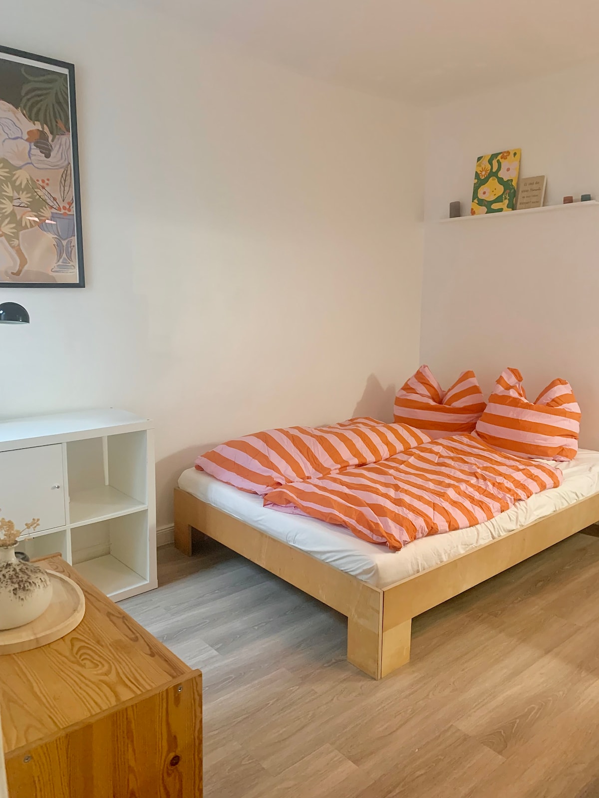 A cozy sleeping area features a spacious bed dressed with orange and white striped bedding. Two accent pillows sit at the head of the bed. A small white shelf is partially visible above the bed, adorned with decorative items. A wooden table with a vase is positioned nearby.