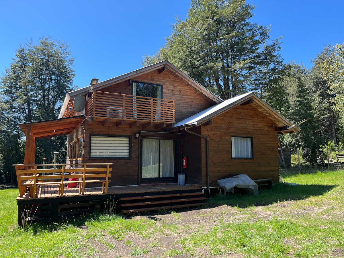 The wooden exterior of the two-story house is complemented by large windows and a balcony. A spacious deck is visible on the lower level, surrounded by a grassy area. Towering trees are seen in the background under a clear blue sky.