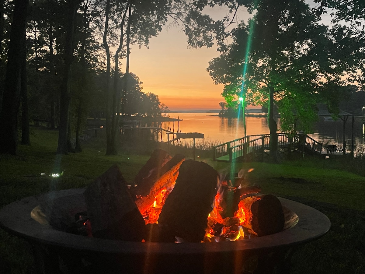 A fire pit filled with glowing embers is surrounded by lush greenery, setting a cozy scene. The lake reflects the soft light of sunset, with silhouettes of trees and a wooden dock visible in the background.