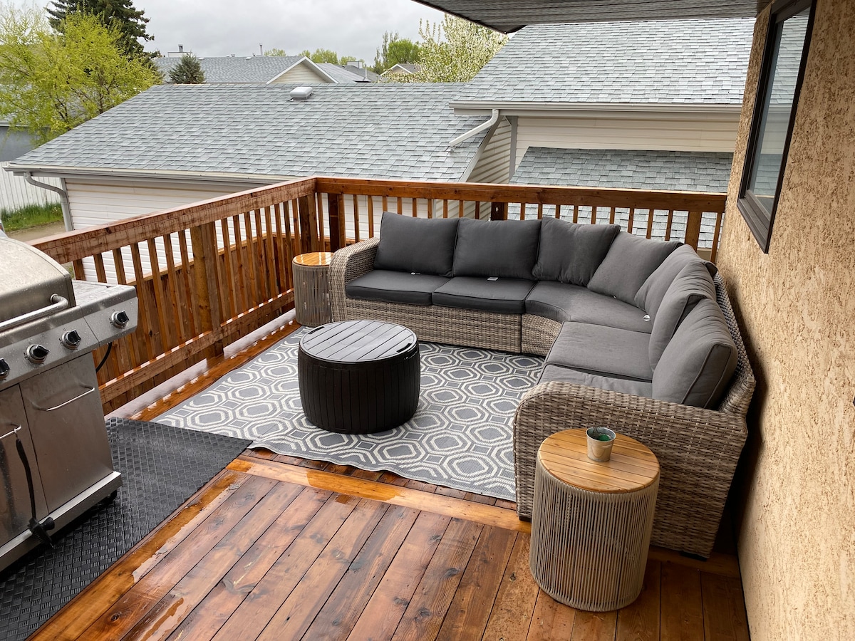 A spacious outdoor seating area is situated on a wooden deck. A comfortable sectional sofa with gray cushions surrounds a circular coffee table. Two side tables are positioned beside the sofa. A grill is visible on the left, with a patterned rug enhancing the space.