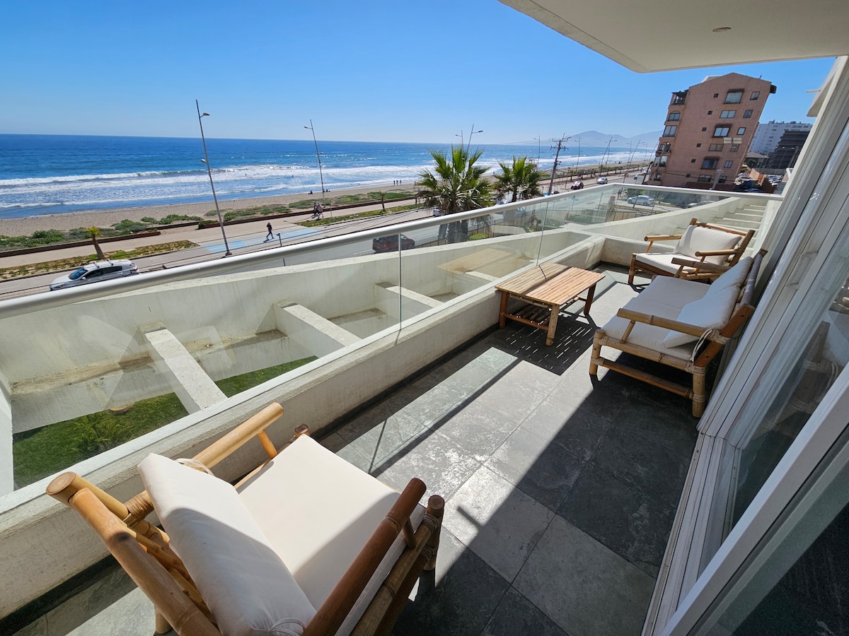 A balcony features two comfortable lounge chairs with soft cushions, overlooking a panoramic view of the ocean. Sunlight reflects off the glass railing, and a small wooden table sits between the chairs. The sound of waves can be heard from this serene outdoor space.