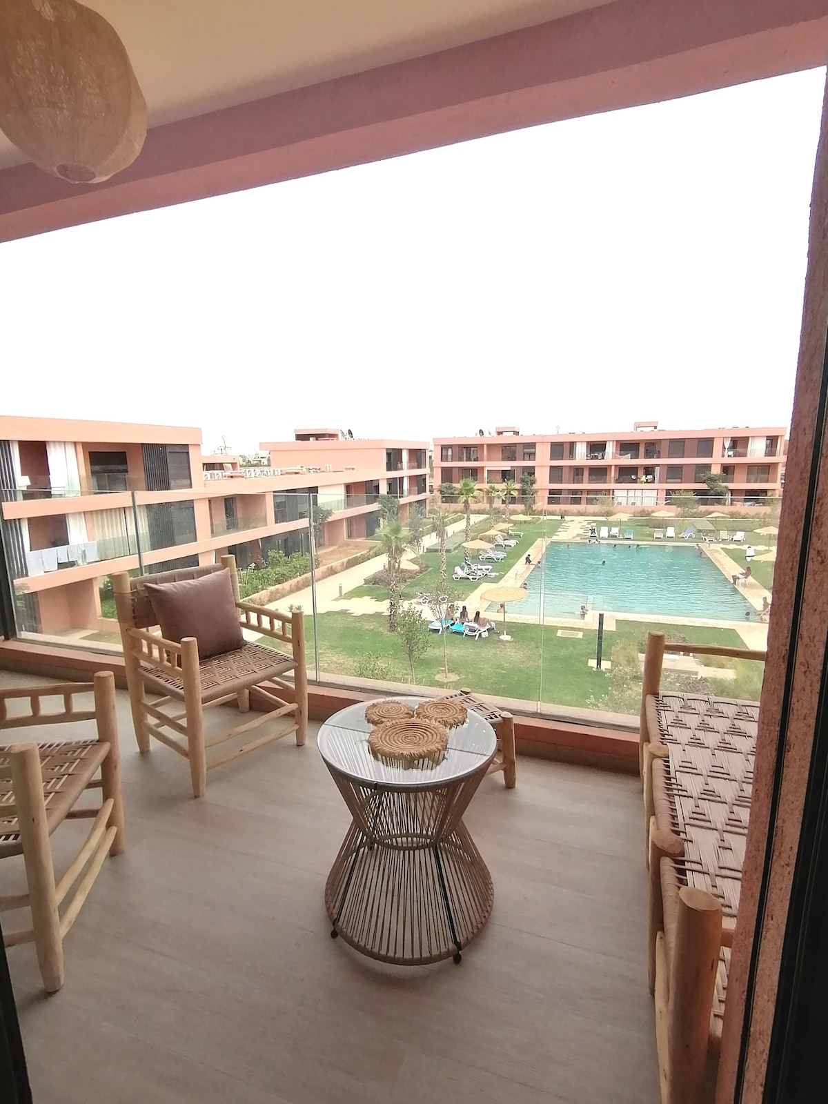 A private balcony is seen, furnished with two bamboo chairs and a round wicker table. The view includes a well-maintained courtyard, featuring a swimming pool surrounded by lounge areas and landscaped gardens. Nearby buildings offer a modern architectural style.