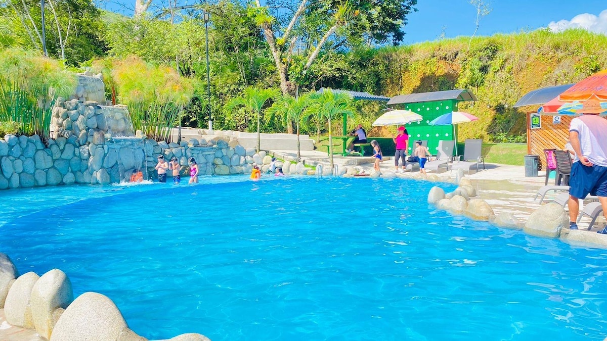 A clear blue pool is surrounded by lush greenery and stone accents. Guests are seen enjoying the water, while shaded lounge areas with colorful umbrellas are visible nearby. The gentle slope of land features a natural stone waterfall, providing a tranquil backdrop.