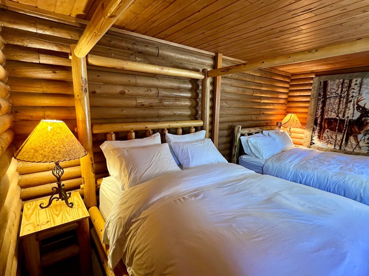 A serene sleeping area features rustic log walls, creating a cozy atmosphere. Two spacious beds are dressed in crisp white linens. A wooden bedside table with a gentle lamp provides soft lighting, complementing the natural warmth of the surroundings.