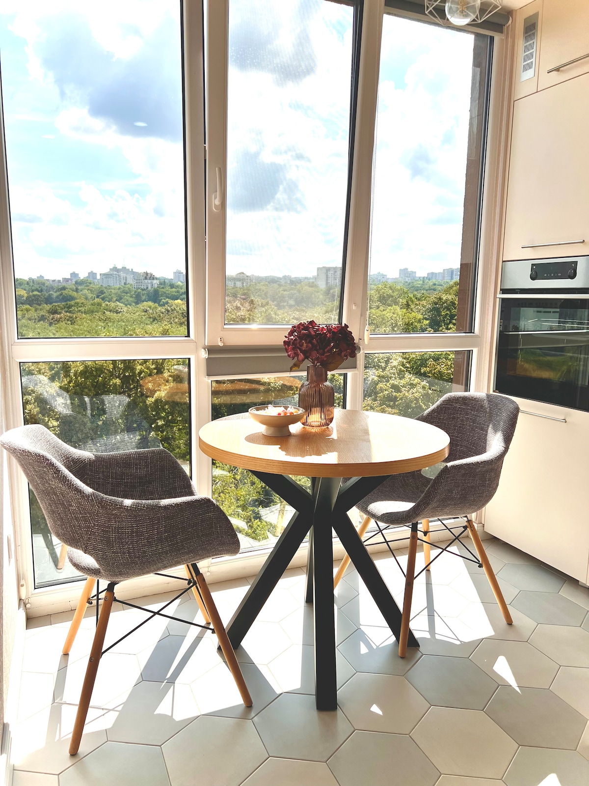 A dining area features a round wooden table set with a delicate vase of flowers. Two grey upholstered chairs are positioned around the table, with large windows providing a view of the lush green landscape outside, allowing natural light to fill the space.