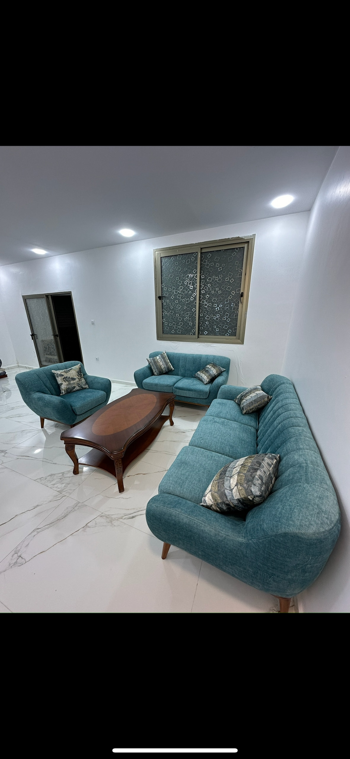 A modern living area features a light blue velvet sofa set positioned around a low wooden coffee table. Two decorative pillows add a touch of comfort to the seating arrangement. A window with a patterned screen allows natural light to illuminate the space.