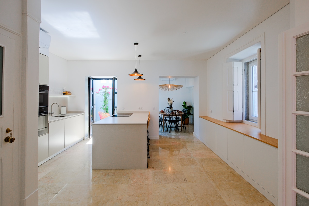 A modern kitchen features sleek white cabinetry and a spacious island with seating. Natural light floods the area through large windows and a door leading to the terrace. The dining area is accessible, showcasing a minimalist design with a wooden table and chairs.