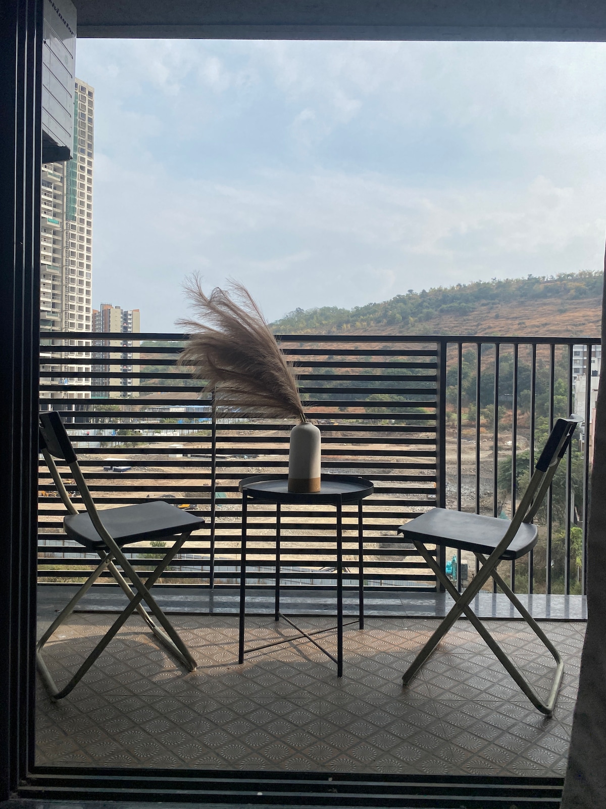A small balcony is equipped with two folding chairs and a circular table. A vase with decorative pampas grass centers the table. A scenic view of distant hills and buildings is visible beyond the railing, with soft natural light illuminating the space.