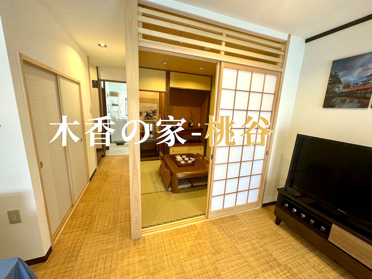A welcoming living area is presented with natural wooden elements and tatami flooring. Shoji screens divide the space, allowing soft light to filter through.  A television is positioned against one wall, complemented by a traditional low table in the center.