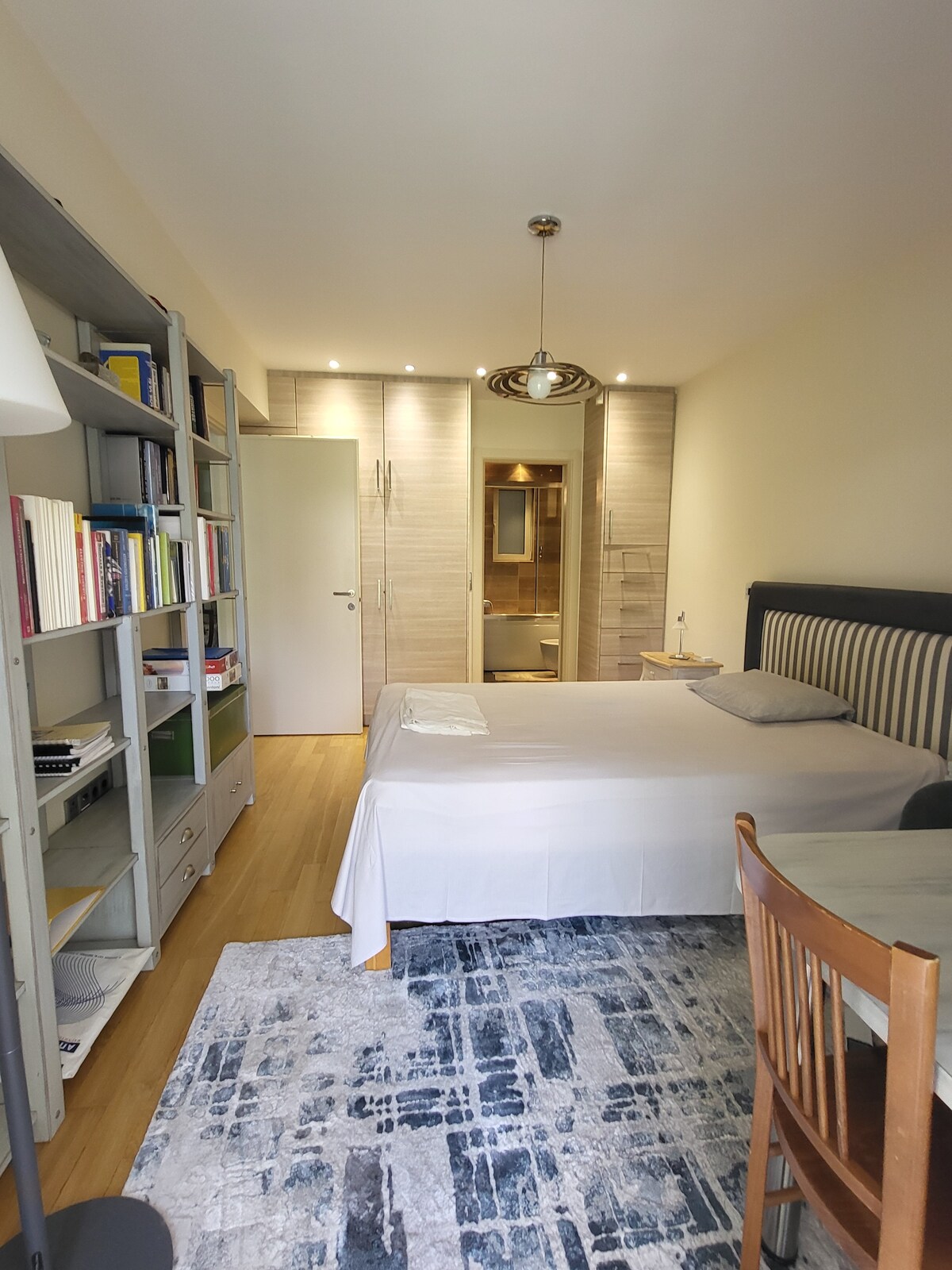 A simple bedroom features a spacious bed draped with a light-colored cover. A wooden chair sits next to a small table, while a bookshelf filled with various books occupies one corner. Soft lighting enhances the warm tones of the room, with modern bathroom facilities visible in the background.