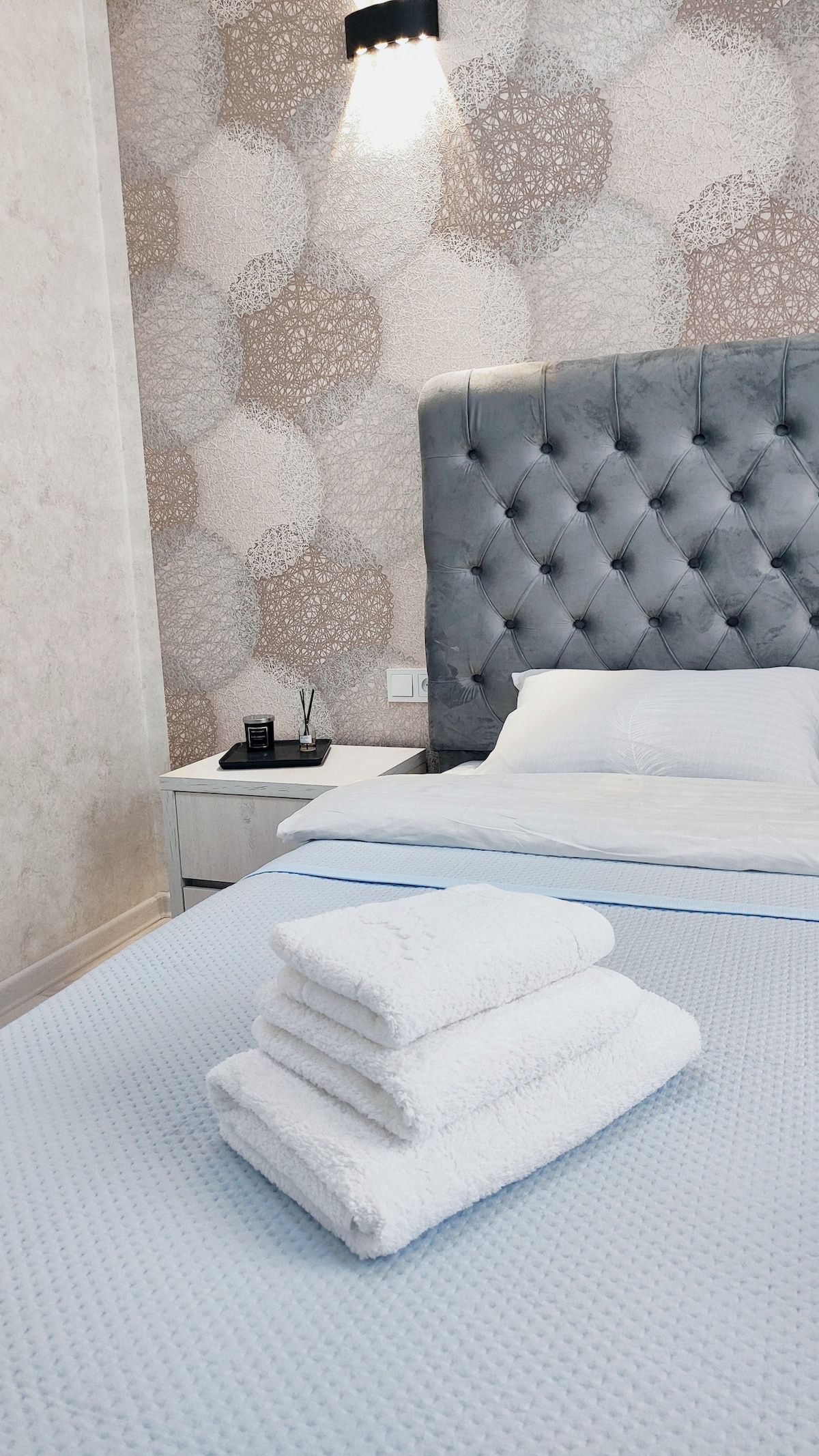 A neatly made bed is presented with soft white towels stacked on top. The headboard features a tufted design, complemented by a light fixture on the wall that enhances the patterned wallpaper in soft tones. A bedside table holds two items, contributing to a minimalist aesthetic.