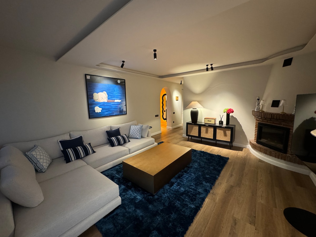 A spacious living area features a light-colored sectional sofa adorned with navy and white cushions. A wooden coffee table is centered on a plush blue rug, and a stone fireplace adds warmth. An artwork depicting gentle waves hangs above, while soft lighting creates a cozy ambiance.