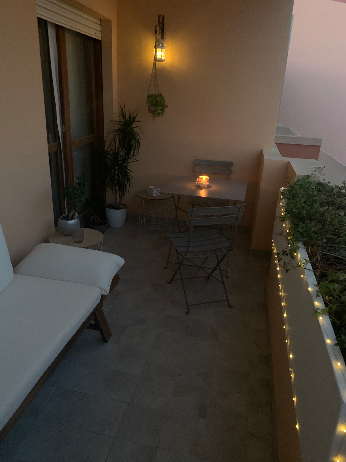 A cozy balcony space features a small table and two chairs, surrounded by plant life. Soft lighting illuminates the area, creating a calm ambiance. A comfortable seating option with cushions is positioned nearby, inviting relaxation in this intimate outdoor setting.