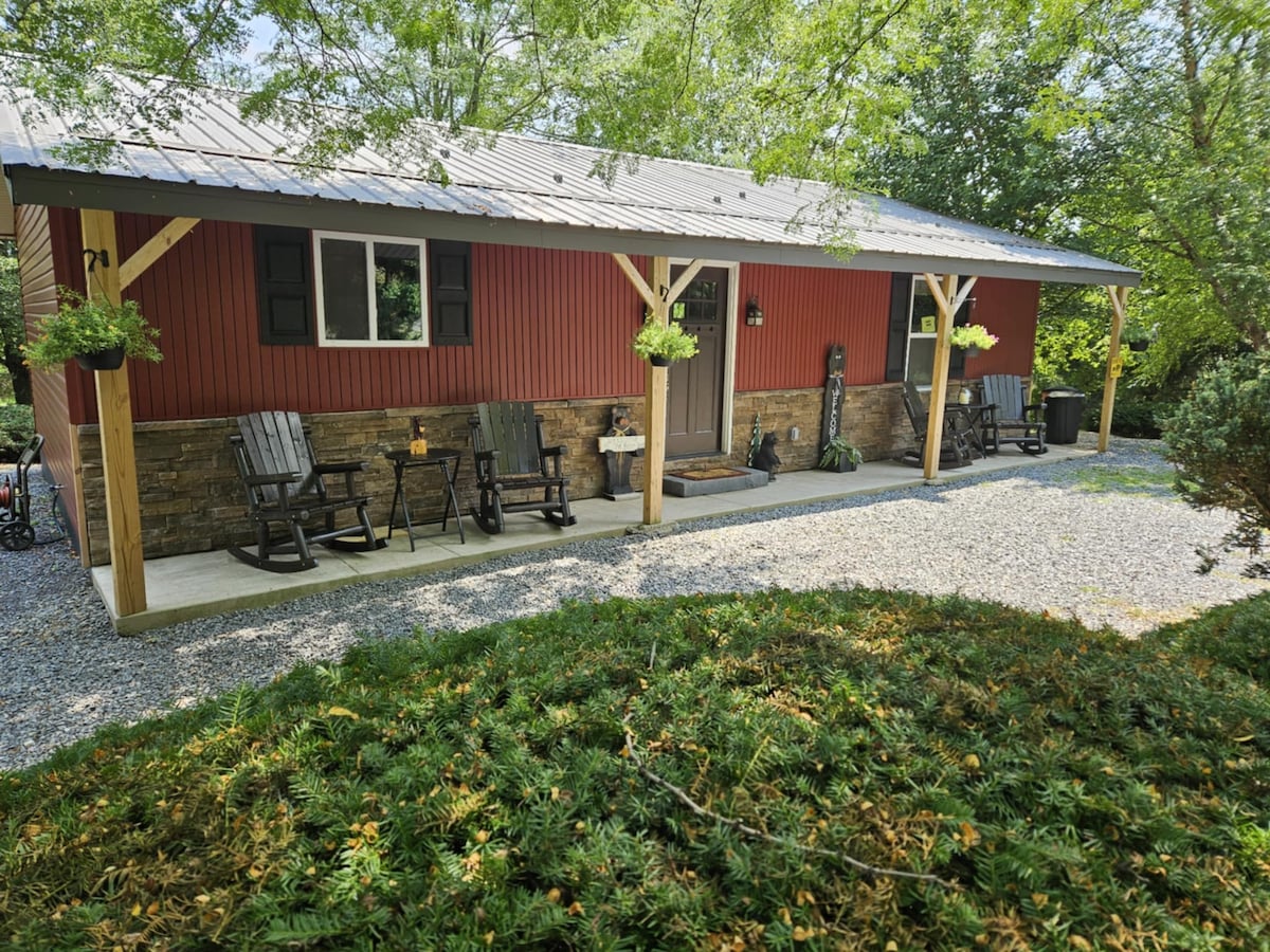 The exterior features a red cottage with a metal roof, surrounded by greenery. A covered porch is supported by wooden beams and includes several rocking chairs. A small table is positioned between the chairs, creating a welcoming entryway, while gravel leads to the entrance.