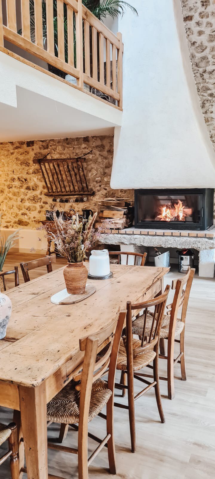 A spacious dining area features a rustic wooden table surrounded by wooden chairs. A large fireplace with a glowing fire serves as a focal point, complemented by natural stone walls. Decorative elements include a vase with dried flowers, adding warmth to the inviting space.