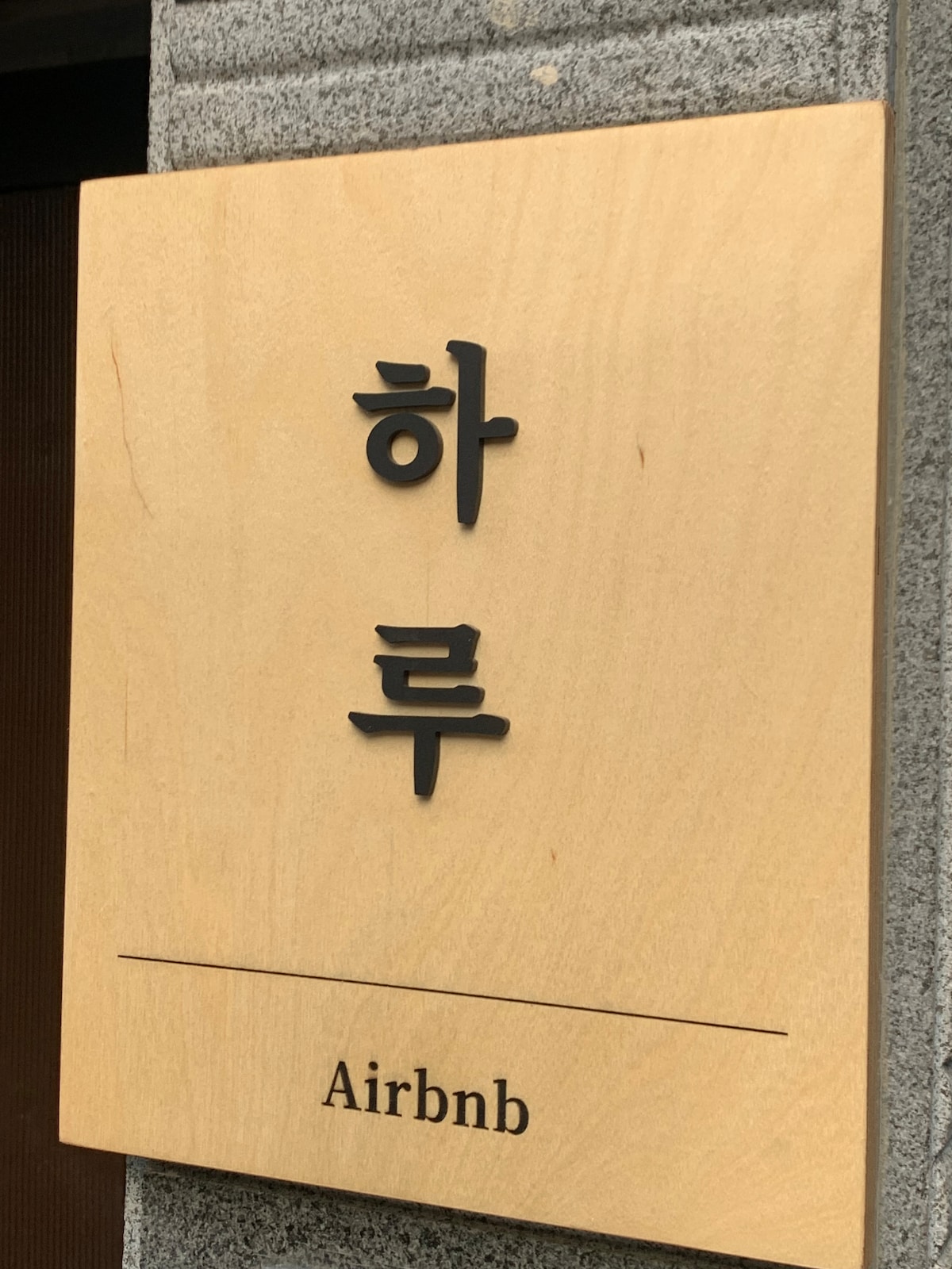 A wooden sign displays the word '하루' in stylish black lettering. Below it, 'Airbnb' is indicated in a subtle, understated font. The sign is affixed to a textured stone wall, contributing to a modern, minimalist entrance.