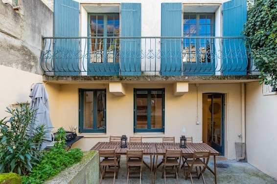 An outdoor dining area is featured with a long wooden table surrounded by several chairs. Two large windows above open to a balcony adorned with blue shutters. Lush greenery frames the space, and a parasol stands tucked in a corner, enhancing the inviting exterior.