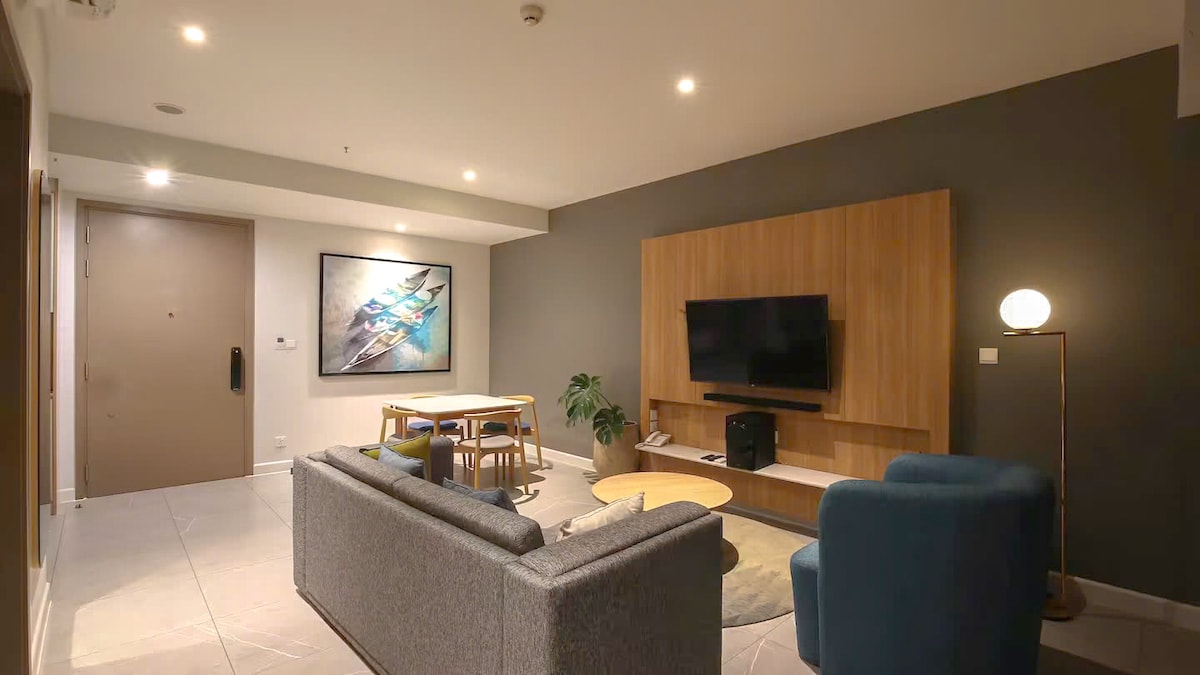 A modern living area features a comfortable sofa, two accent chairs, and a dining table set for four. A large flat-screen TV is mounted on a wooden wall unit. A piece of artwork adorns the wall, adding a pop of color to the neutral decor.