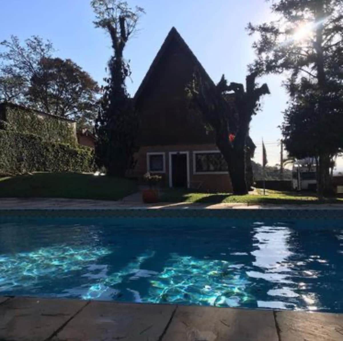 A serene swimming pool reflects sunlight, with a charming sloped-roof house visible in the background. Lush trees and greenery surround the area, creating a tranquil outdoor setting perfect for relaxation.