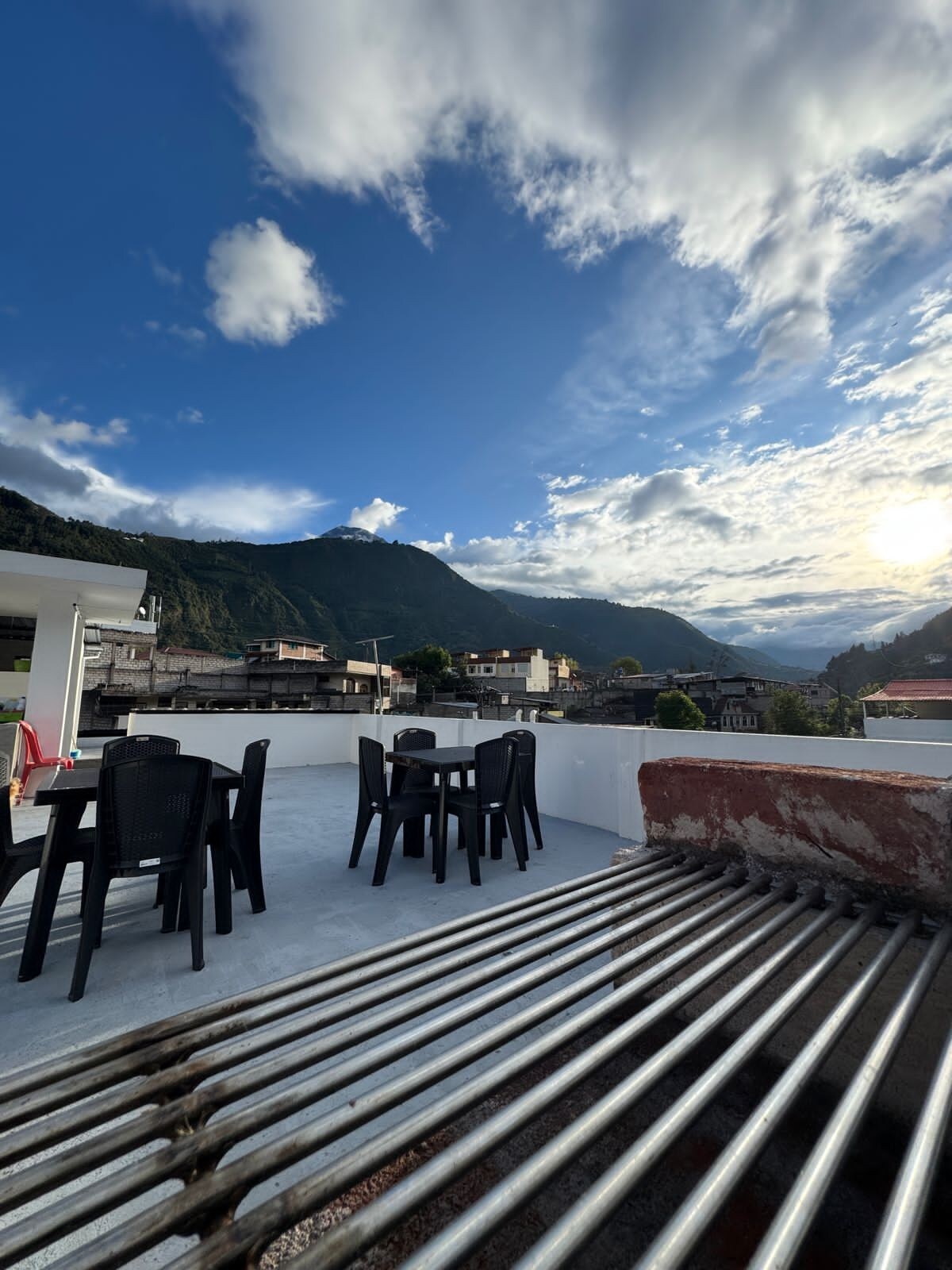 A spacious rooftop area features several black plastic chairs and tables. Expansive views of the surrounding mountains and the sky, characterized by scattered clouds and sunlight, enhance the serene outdoor atmosphere.
