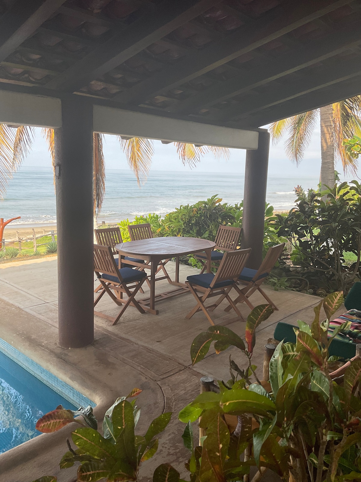 A shaded outdoor dining area is furnished with a wooden table surrounded by six folding chairs. The setting offers views of the ocean through the palm trees, complemented by a glimpse of the infinity pool nearby. Tropical plants line the edge of the space.