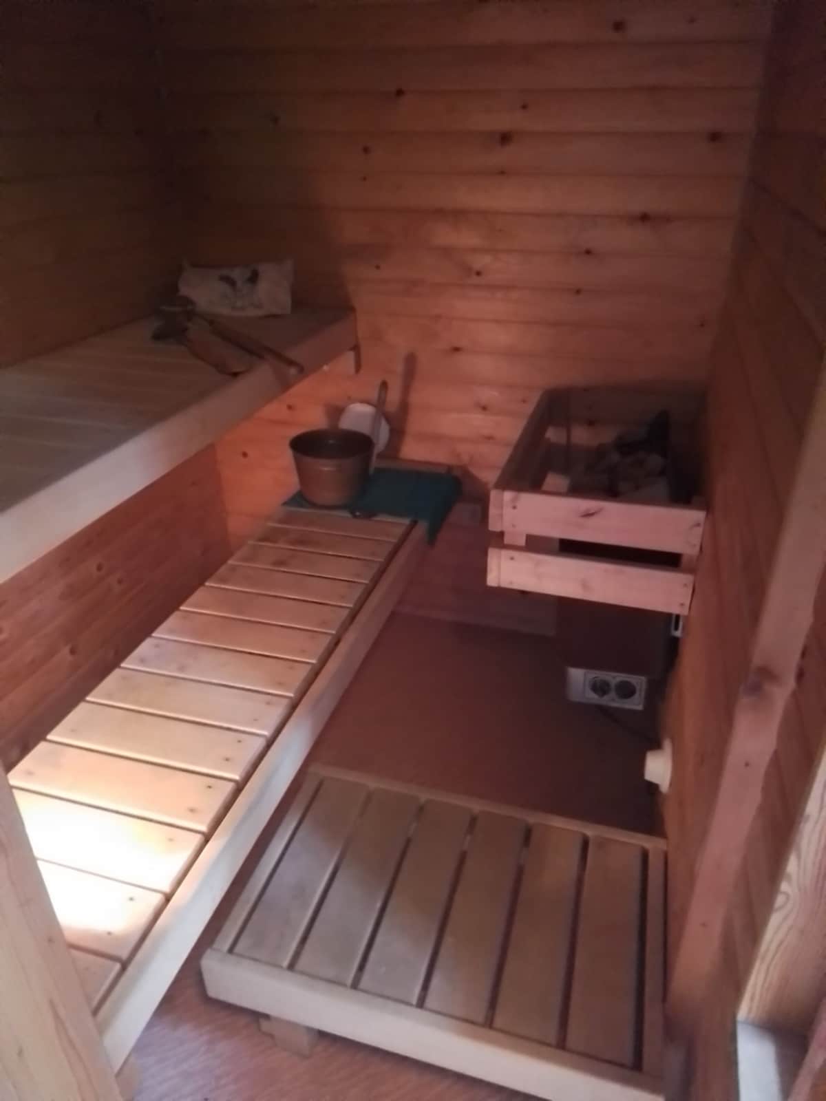 A wooden sauna interior features clean lines with a bench along the left wall and an elevated platform. A bucket and ladle are placed on a small table, with another bench section located opposite. The warm wood tones create a serene retreat for relaxation.