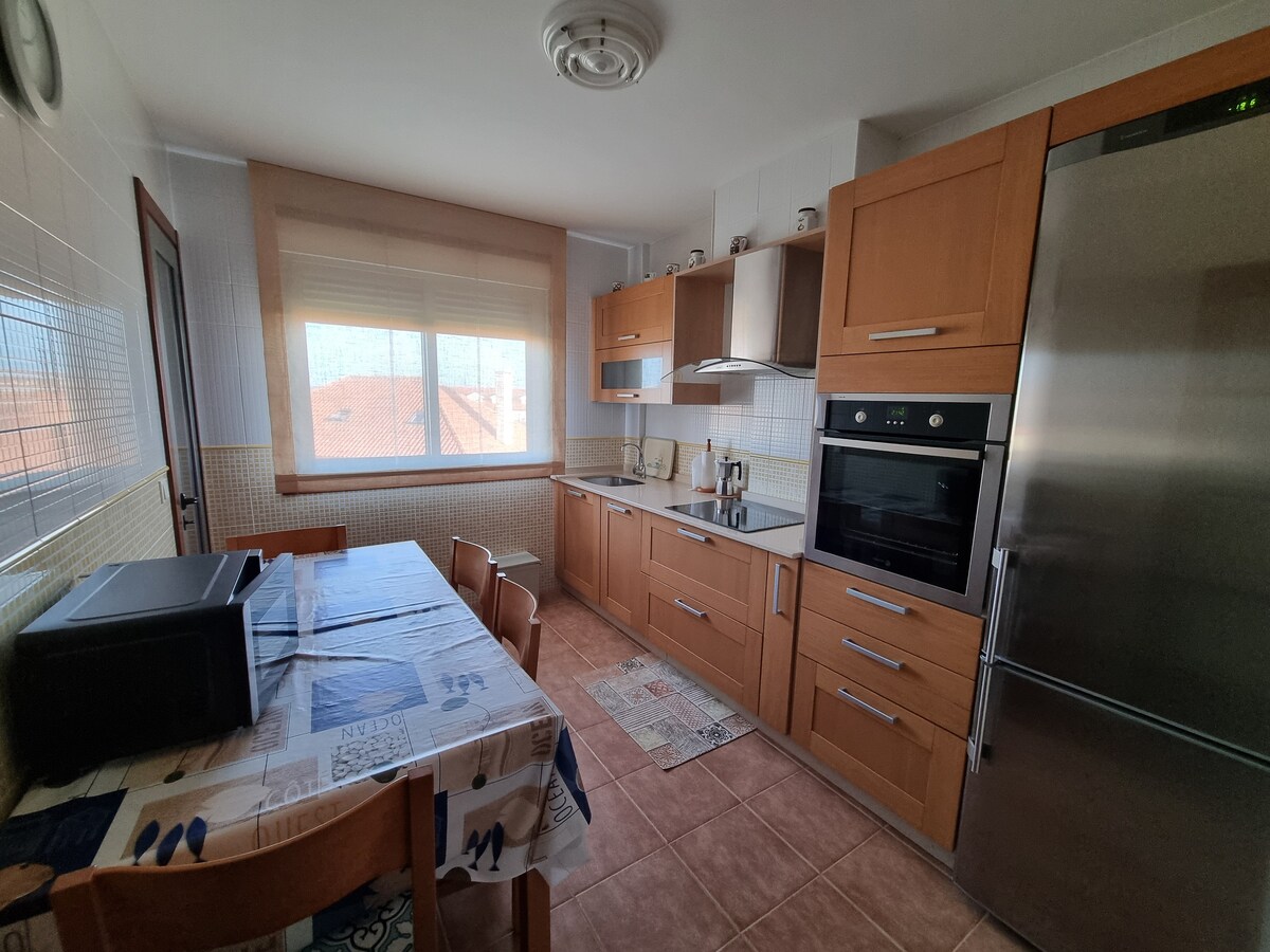 A well-equipped kitchen is presented, featuring wooden cabinetry and modern appliances. A dining table with wooden chairs occupies one area, complemented by a light-colored rug nearby. Natural light enters through a large window, enhancing the space's openness.
