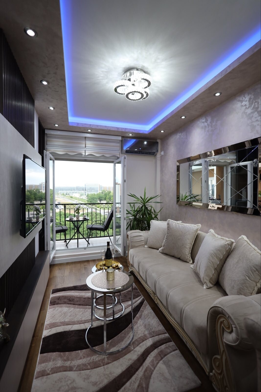 A cozy living area features a light-colored sofa adorned with decorative pillows. A round glass table is positioned on an elegant area rug. Bright ceiling lights and blue accent lighting create a soft ambiance. French doors lead to a balcony with views of greenery.