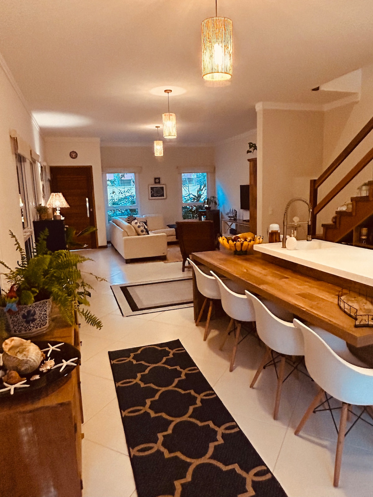The interior space features an open layout with a dining area showcasing a wooden table surrounded by white chairs. A comfortable seating area is visible, accompanied by decorative plants and soft lighting from ceiling fixtures. A staircase leads to the upper level, providing access to additional rooms.