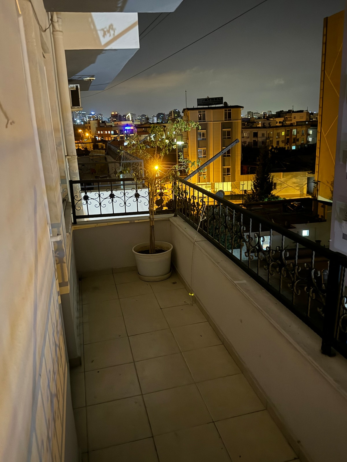 A private balcony is featured, surrounded by a wrought iron railing. A potted plant adds a touch of greenery. The ground is tiled, and city lights twinkle in the background against a night sky.