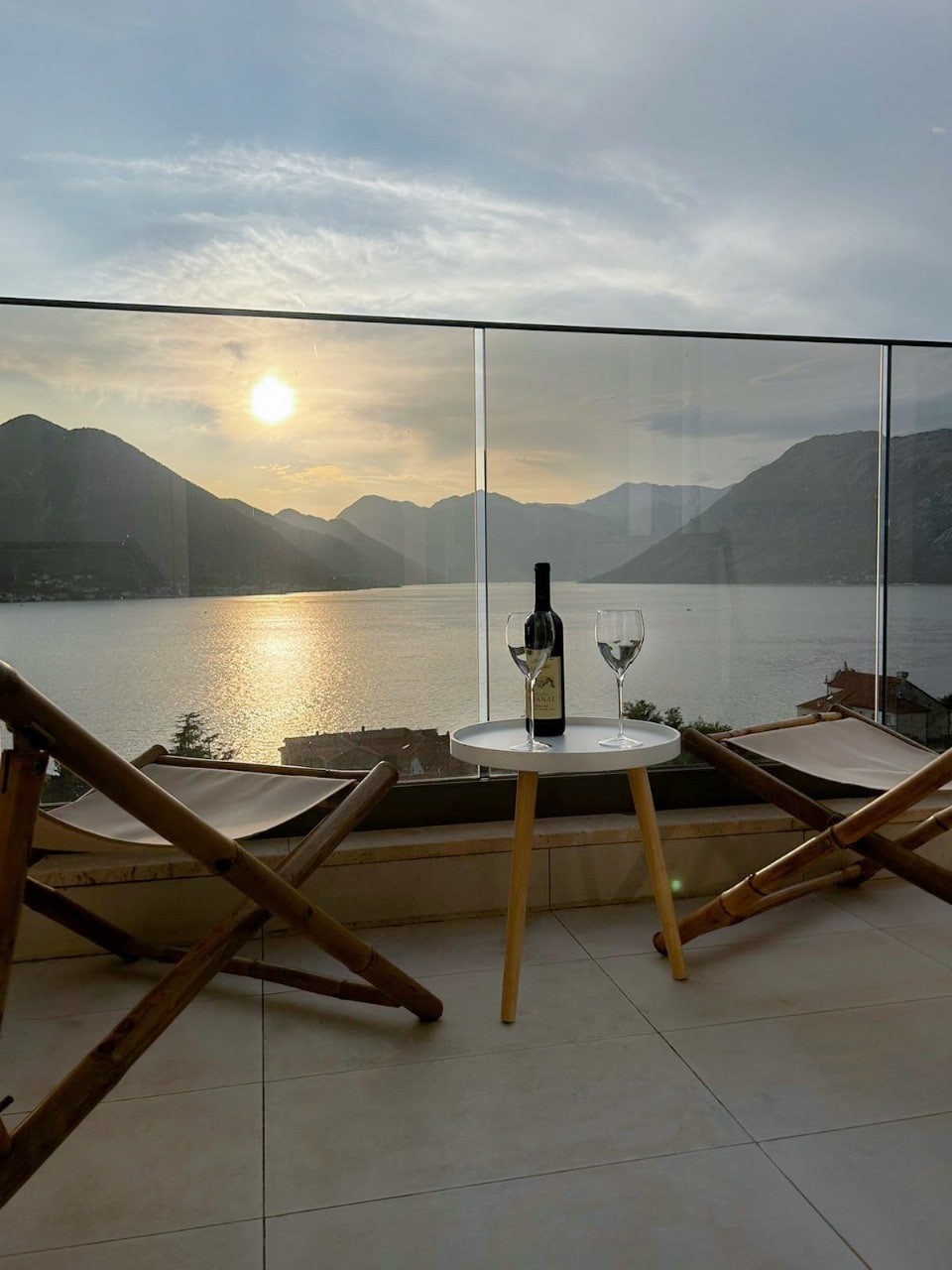 A serene outdoor balcony features two wooden lounge chairs positioned towards the stunning view of mountains and a calm sea at sunset. A small round table holds a bottle of wine and two glasses, enhancing the relaxing atmosphere.
