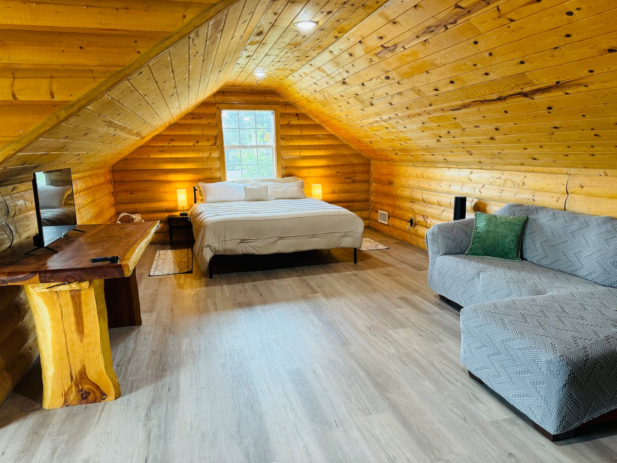 An open loft space is showcased with sloped wooden ceilings and warm wooden walls. A spacious bed is positioned against a window, while a cozy sofa is placed nearby. A wooden table offers a flat-screen TV, contributing to a comfortable and inviting atmosphere.