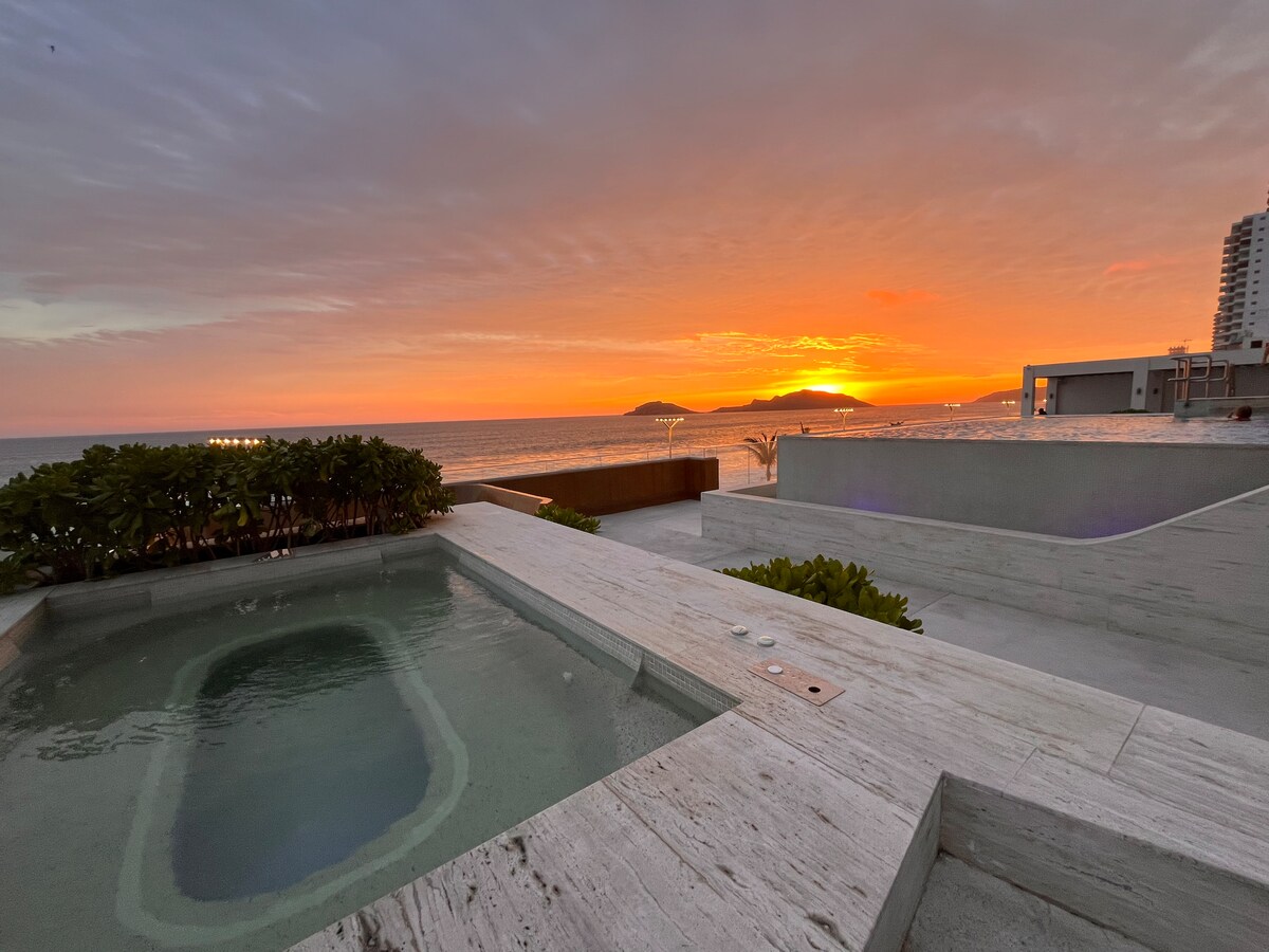 A tranquil outdoor area is highlighted by a jacuzzi surrounded by smooth stone surfaces. The view features a vibrant sunset over the ocean, with gentle waves visible in the distance. Lush greenery lines the space, enhancing the relaxed atmosphere.