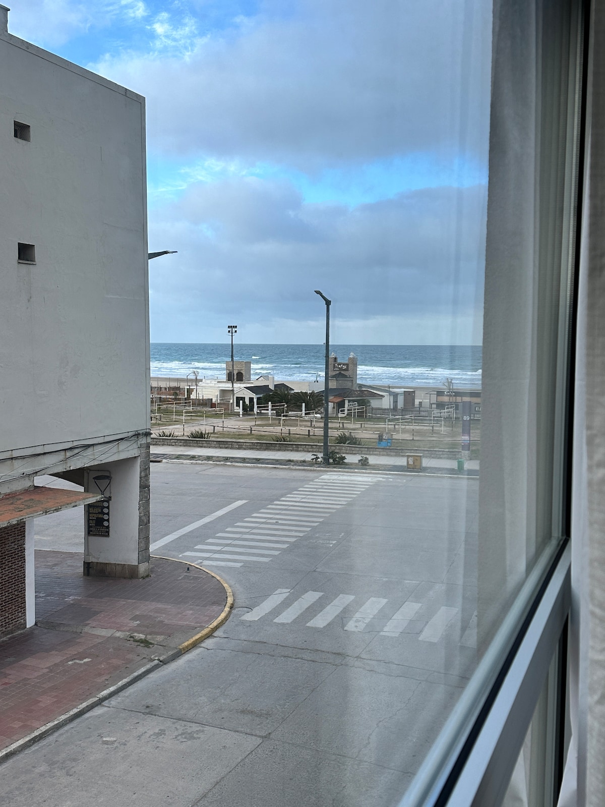 A view from a window showcases the ocean with rolling waves under a partly cloudy sky. The scene includes a street with a crosswalk and streetlights, along with a glimpse of a beachfront area featuring trees and grassy spaces.
