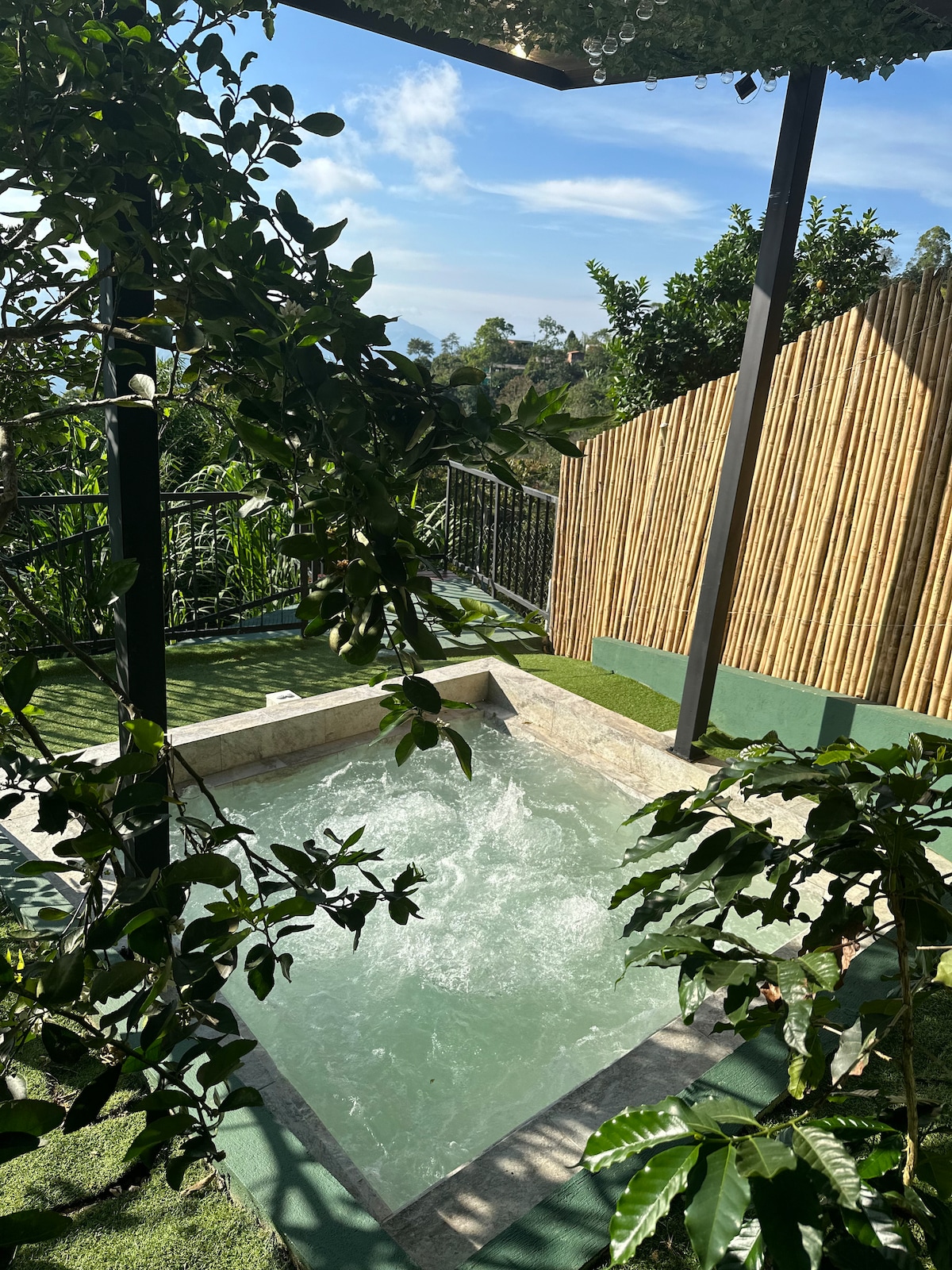 A serene outdoor jacuzzi is surrounded by lush greenery, offering a peaceful retreat. The bubbling water reflects the sunlight, while a bamboo fence provides privacy. The area is designed for relaxation, with a gentle slope leading towards the scenic landscape visible in the background.