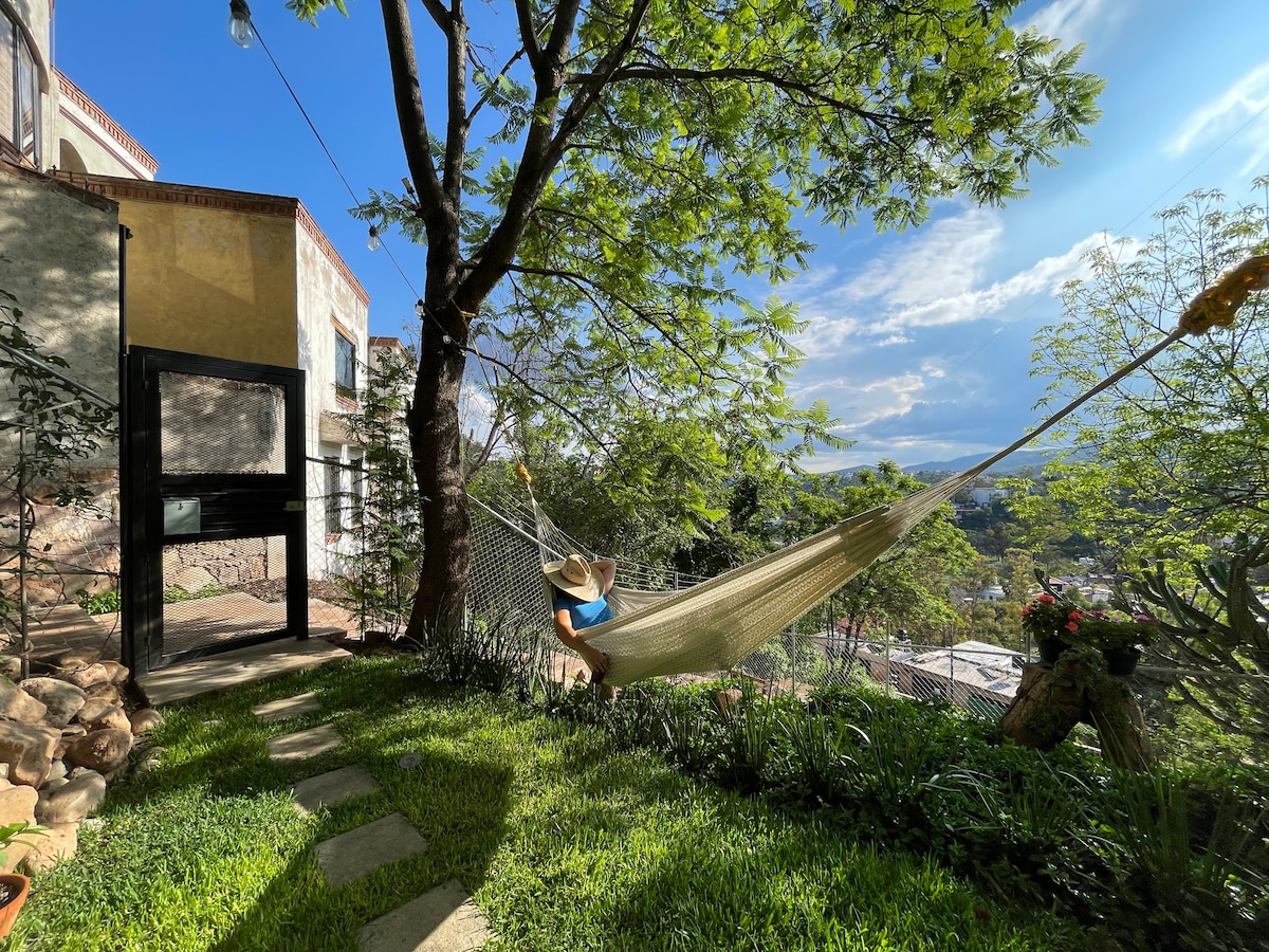 A hammock is suspended between two trees in a lush green garden. A pathway of stepping stones leads through the grass, with gentle sunlight illuminating the scene. A panoramic view of the city is visible in the background, enhancing the serene outdoor space.