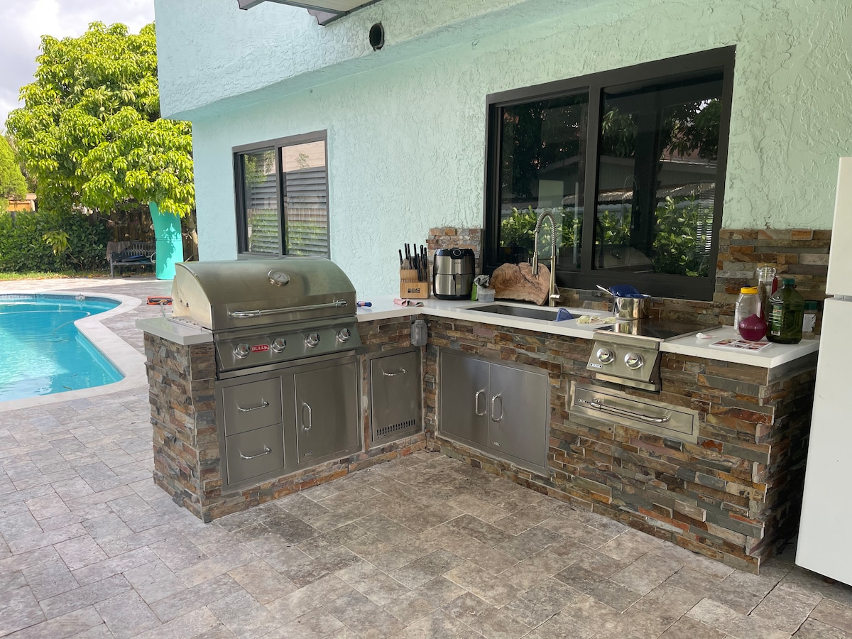 An outdoor kitchen area is featured next to a pool, showcasing a grill integrated into stone cabinetry. A countertop is equipped with various utensils and a sink, with greenery visible beyond the window. The surrounding space is paved and offers a seamless transition to the pool area.