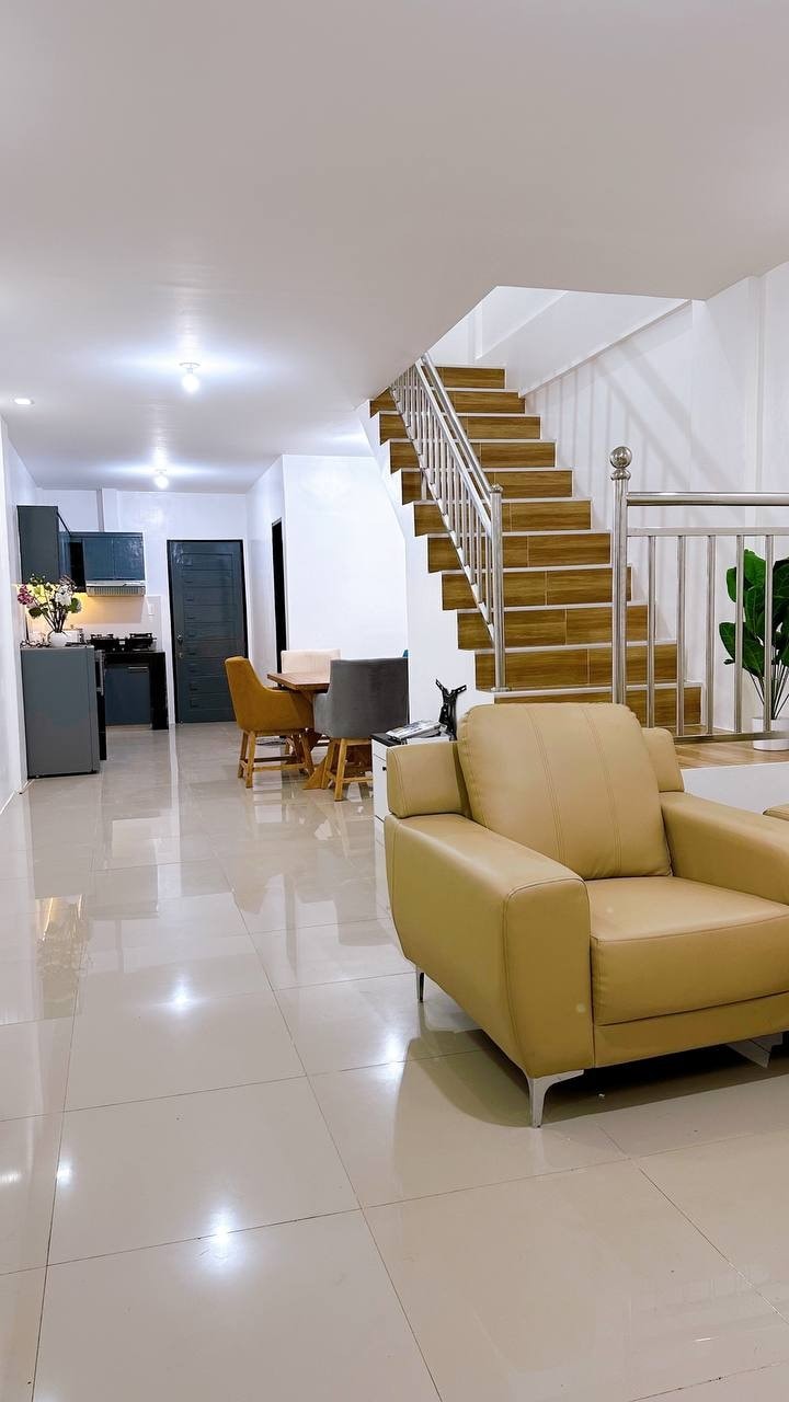 A spacious living area is showcased, featuring a light-colored armchair and a staircase leading to the upper level. The floor is tiled in a reflective finish, enhancing the airy feel. A dining area with chairs is visible in the background, adjacent to an open kitchen space.