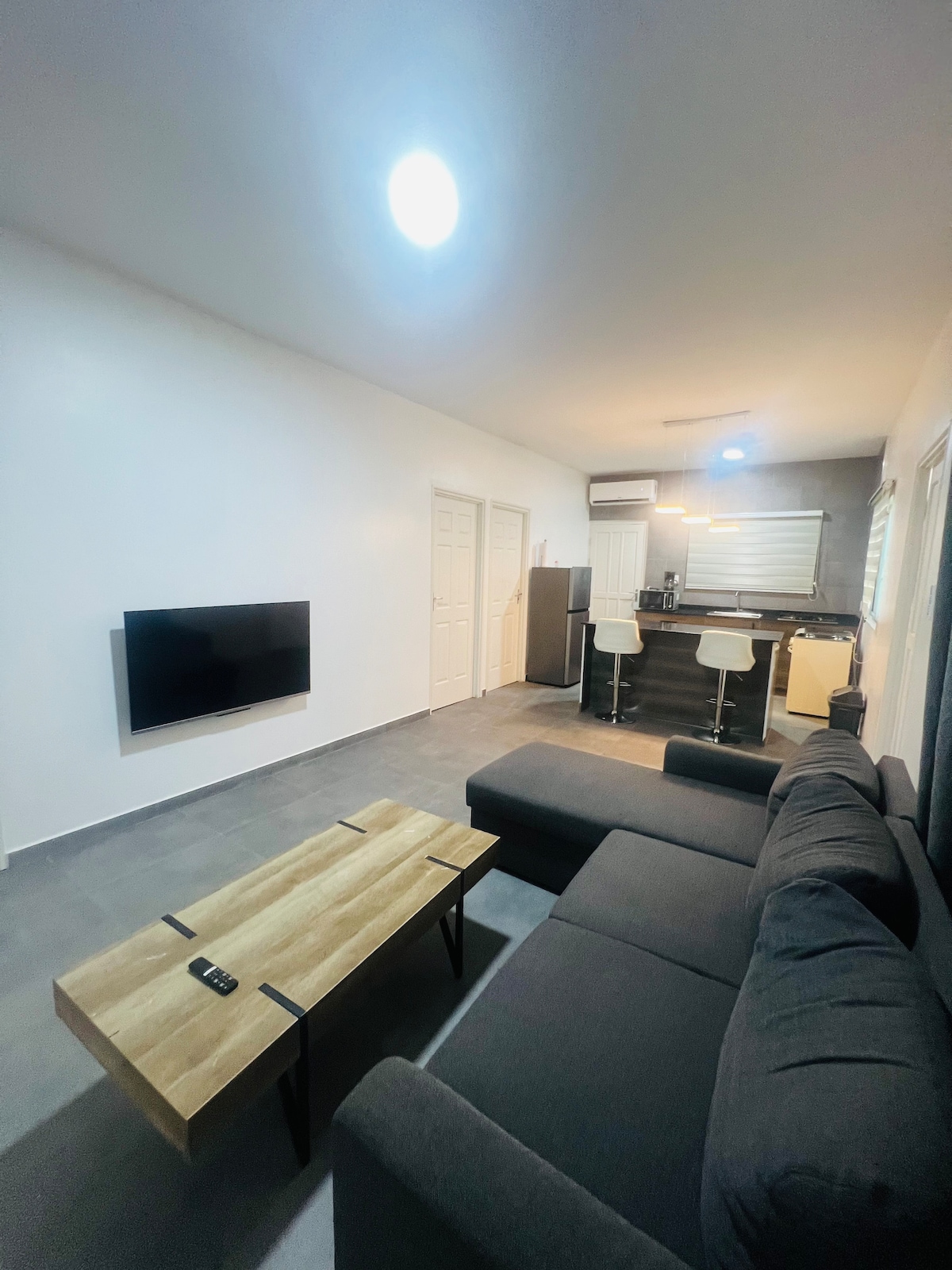 A modern living area features a dark sectional sofa positioned around a wooden coffee table. A wall-mounted television is visible across from the seating. An open kitchen is partially seen in the background, equipped with minimalist bar stools and a light fixture above.