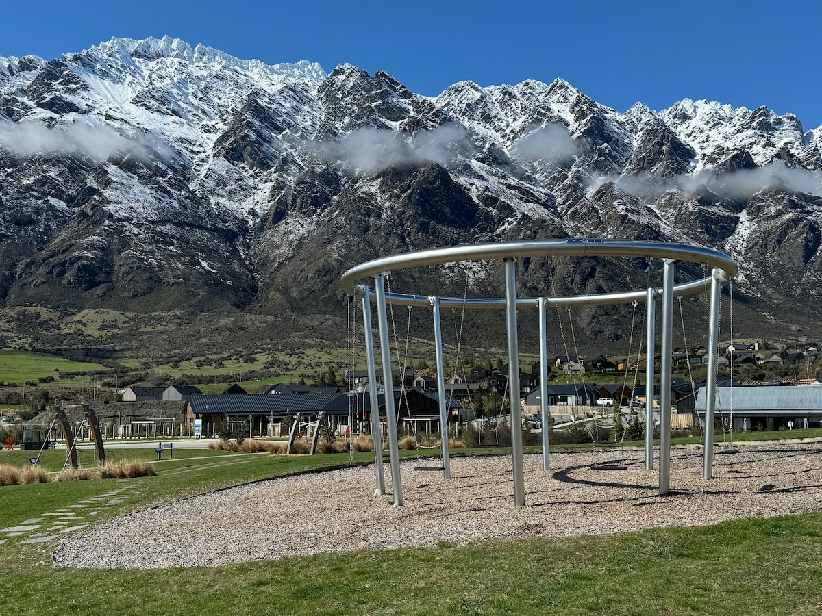A modern playground structure is featured in the foreground, surrounded by a gravel area. The backdrop showcases a dramatic mountain range covered in snow, with blue skies above. The scene includes green grass and distant buildings visible in the peaceful landscape.